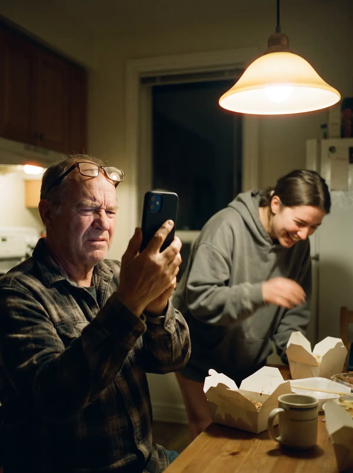 A dad squints at his new phone at arm’s length, reading glasses pushed up on his forehead, while his daughter doubles over laughing behind him