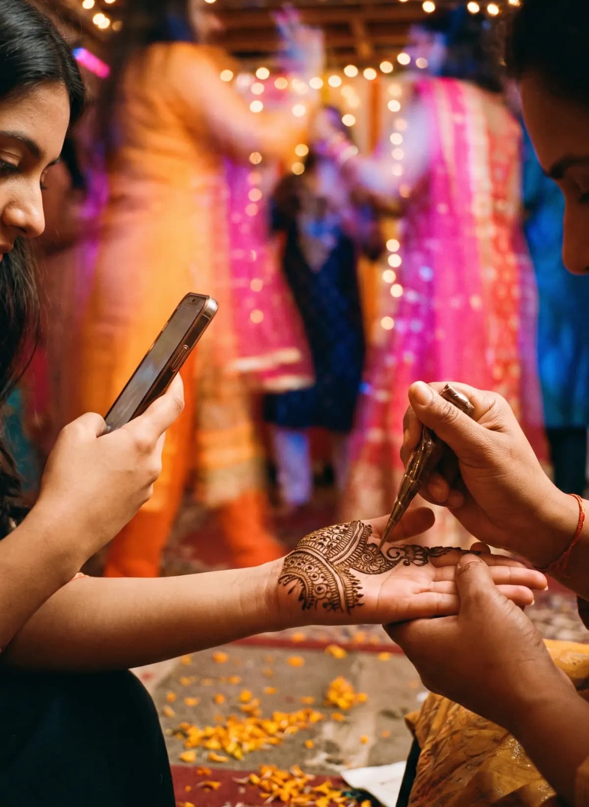 A henna artist works on a palm while the bride’s sister films it and cousins dance in a blur behind