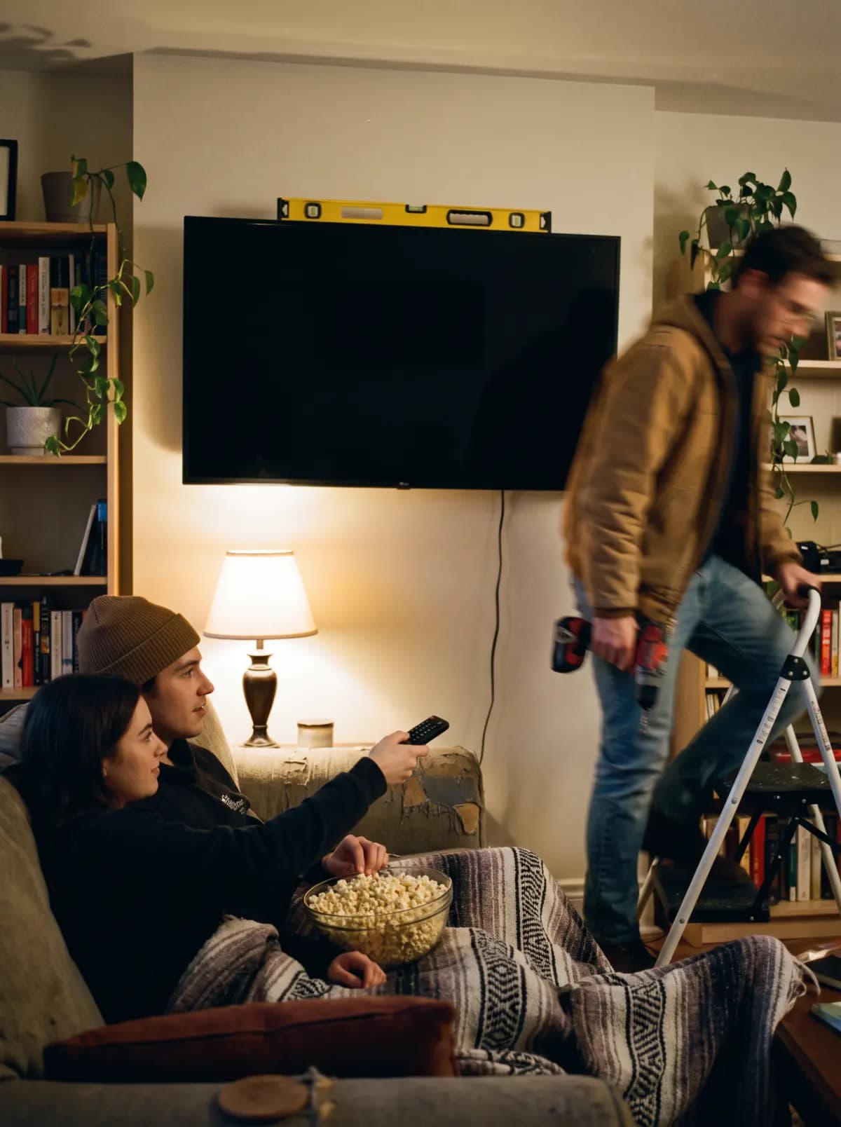 A TV freshly mounted with no cables showing as the handyman steps down and a couple already waits with popcorn