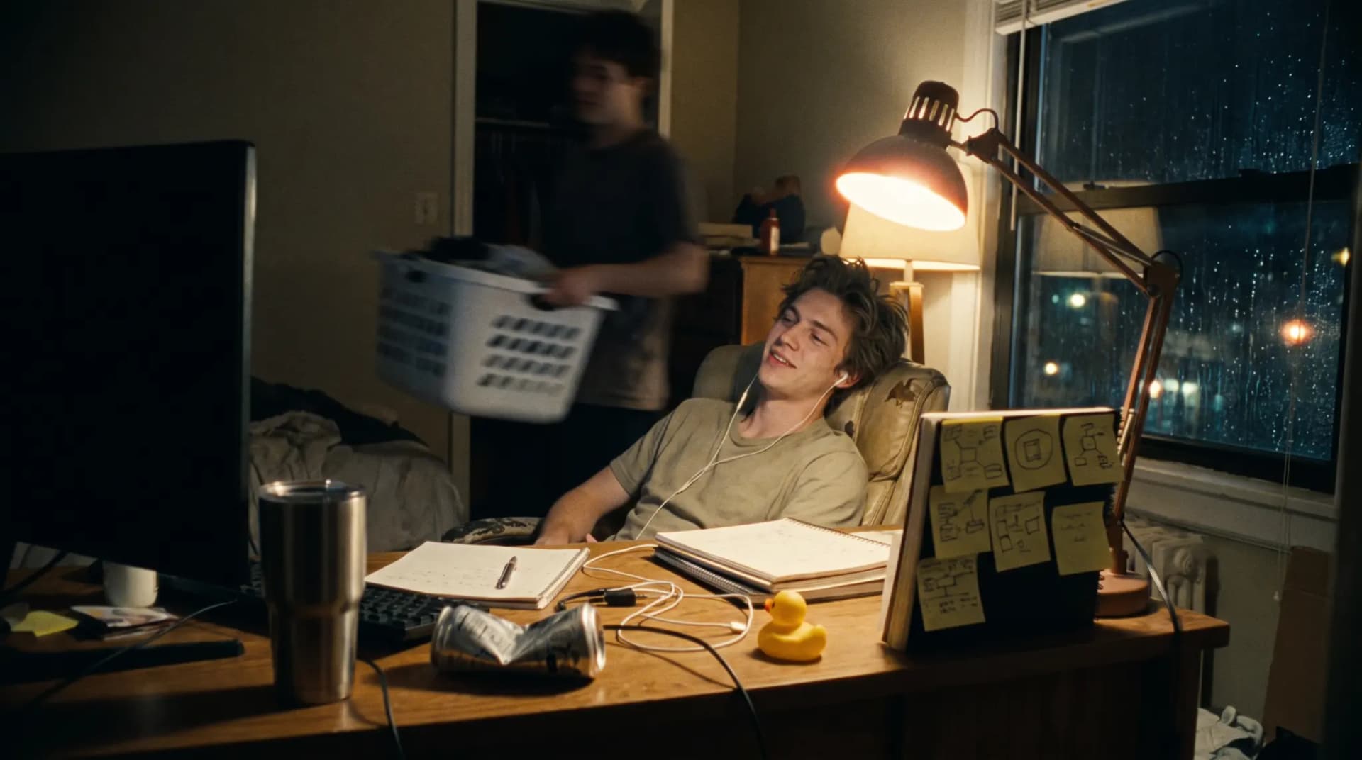 A young engineer at a desk late at night leans back with a half-smile as something finally works, chai and a rubber duck by the keyboard, a housemate blurred behind