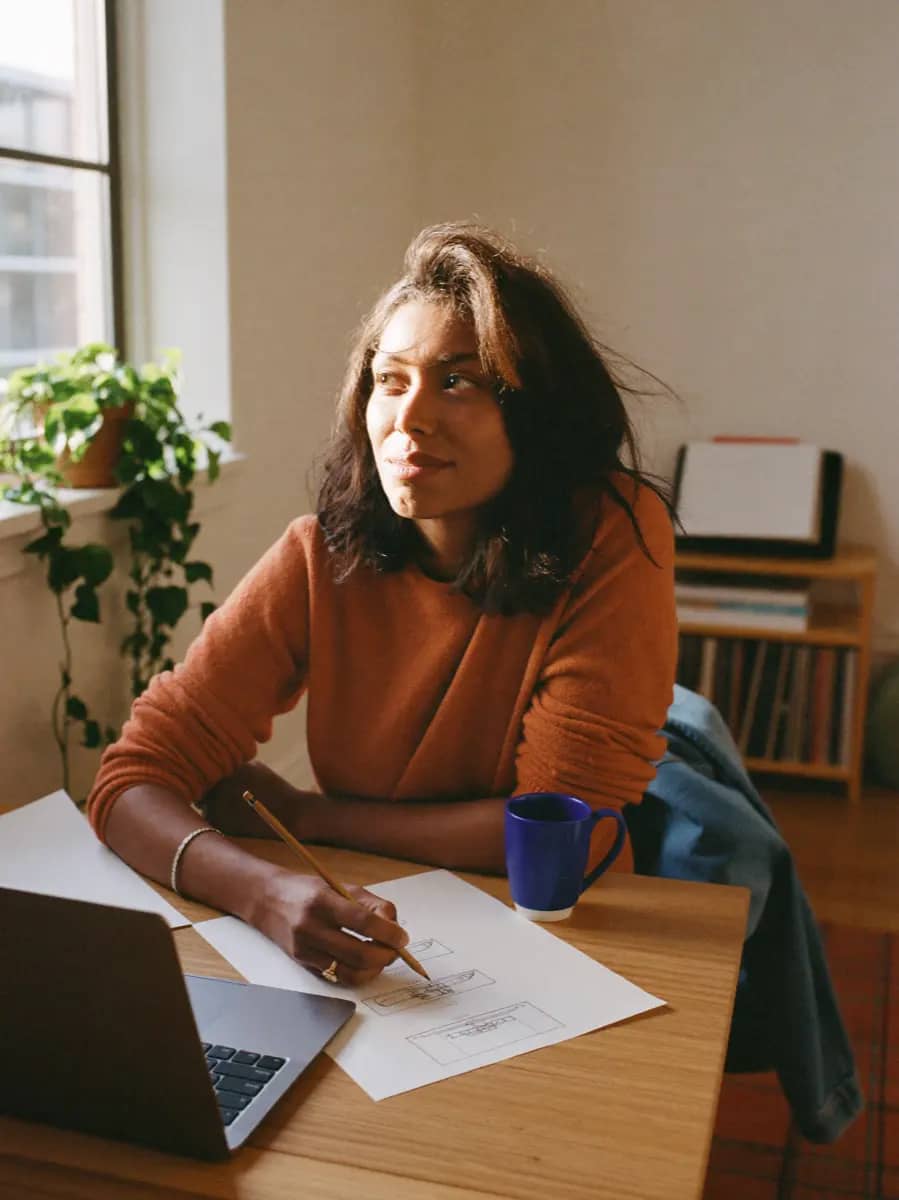 A designer glancing up from pencil thumbnails beside an open laptop in a sunlit home studio.