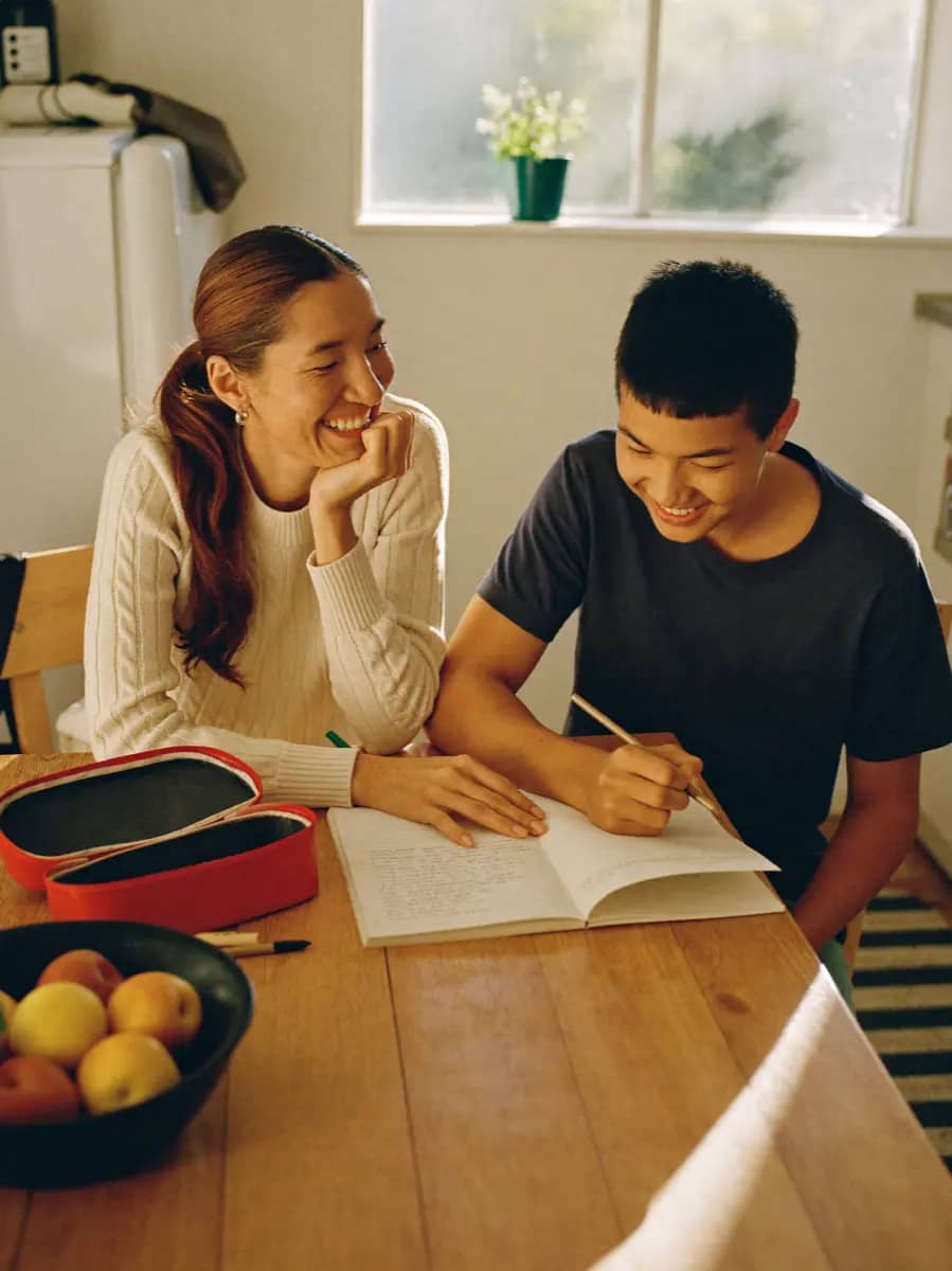 A tutor laughing with a student over an open notebook at a kitchen table.